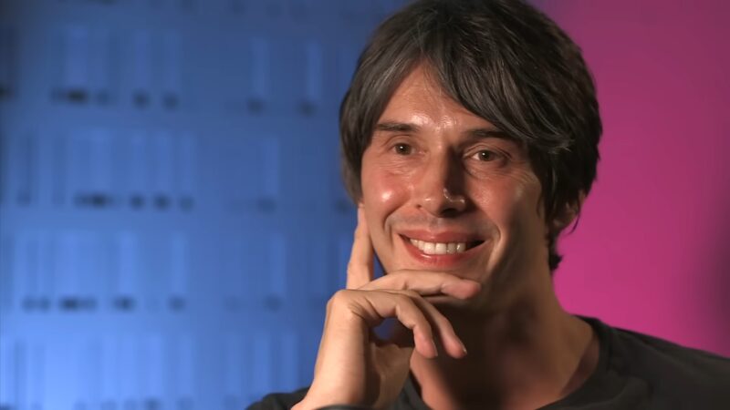 Brian Cox smiling during a studio interview with a blurred science-themed background