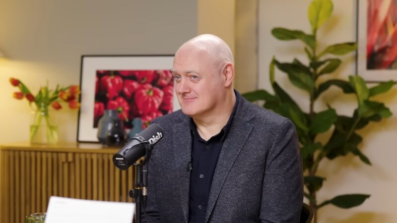 Dara Ó Briain sitting at a table with a microphone during a studio interview