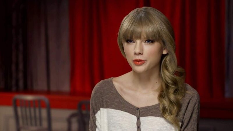 Blonde female singer, Taylor Swift, speaking in front of red curtains.
